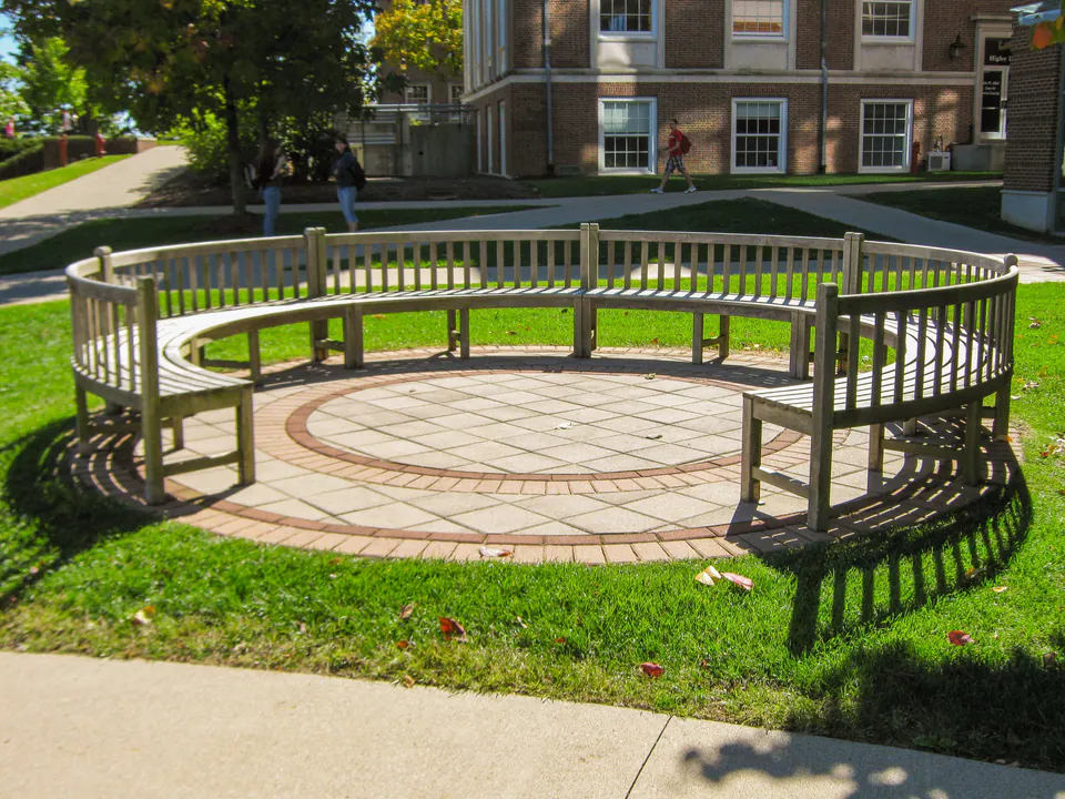Circular campus bench plaza with curved wood seating, paver circle, lawn, and walkway edge